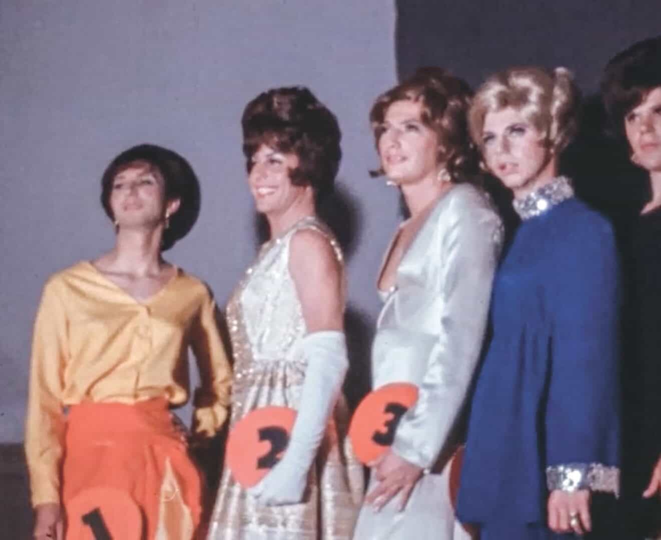 Five women in vintage dresses and costumes on stage with numbers 1-4.