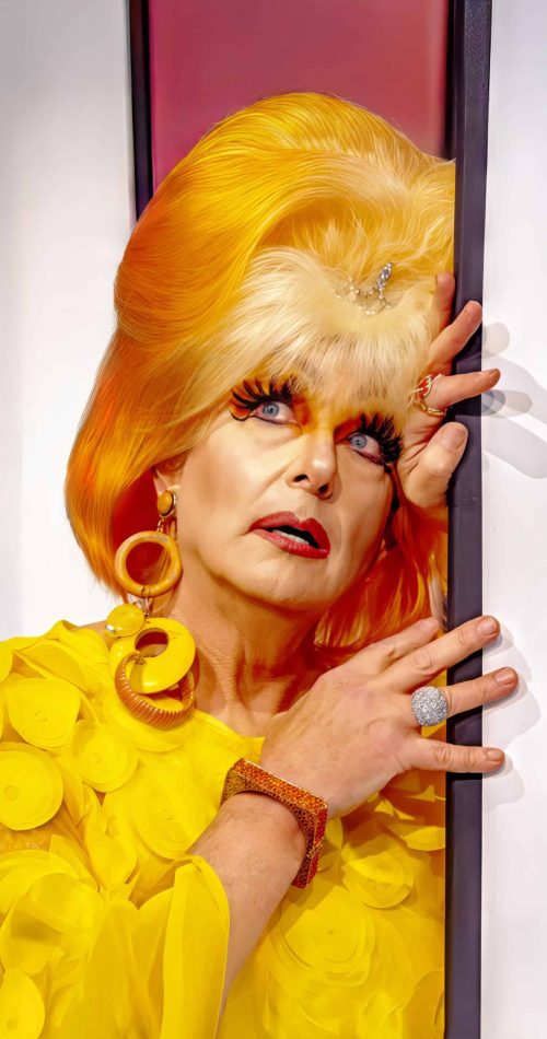 Performer in feathered gown and bright yellow hair peeks through a door. — friendsofgeorges.org